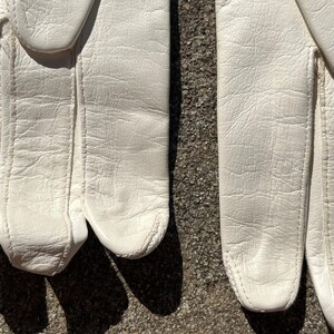 May include: Close-up of a pair of white leather gloves. The gloves are made of a textured leather material and have visible stitching along the seams. The gloves are lying on a rough, gray surface, with shadows cast across the surface.