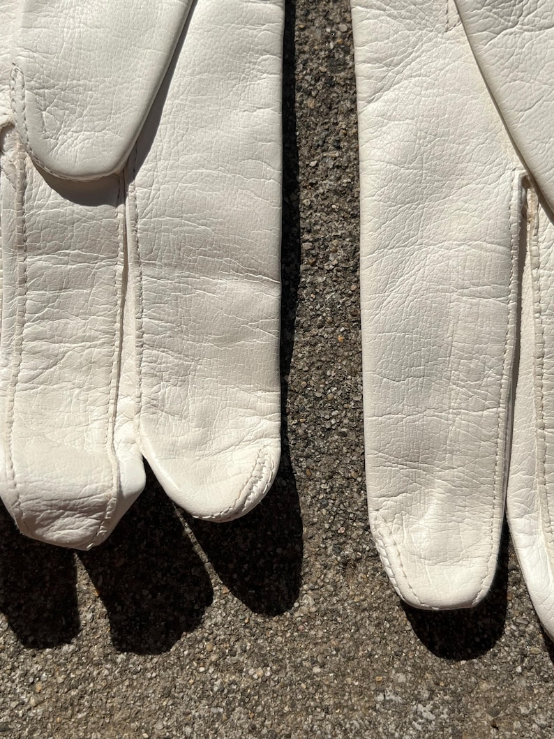 May include: Close-up of a pair of white leather gloves. The gloves are made of a textured leather material and have visible stitching along the seams. The gloves are lying on a rough, gray surface, with shadows cast across the surface.