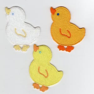 Op de afbeelding: Drie vilten kuikenpatches in wit, geel en oranje. Elk kuiken heeft een snavel, een oog en voetjes in contrasterende kleuren. De patches zijn waarschijnlijk voor knutselwerkjes of versieringen, perfect voor Pasen of projecten met een lentethema.
