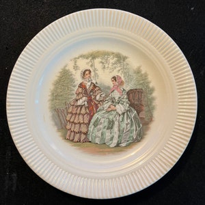 May include: A white ceramic plate with a raised rim and a floral design featuring two women in 19th-century dresses. The women are standing in a garden setting with trees and bushes.