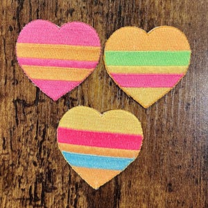 May include: Three heart-shaped patches with horizontal stripes in vibrant colors. One heart features pink and orange stripes, another has orange, green, and pink stripes, and the third showcases yellow, pink, blue, and orange stripes. The patches are on a wooden surface.