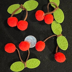 Puede incluir: Un conjunto de cerezas de fieltro con hojas de fieltro verde unidas a cordones marrones. Las cerezas son rojas y las hojas son verdes. Las cerezas están dispuestas en un racimo sobre un fondo negro.