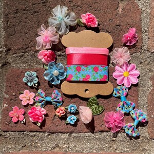 Può includere: Una collezione di materiali per l'artigianato tra cui nastri rosa, rossi e blu, insieme a vari abbellimenti floreali. I nastri sono esposti su una bobina marrone, e i fiori e i fiocchi sono in tonalità di rosa, blu e bianco.
