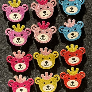 Op de afbeelding: Twaalf glitzerende teddybeer-knoppen met kronen. De beren zijn roze, blauw, geel en rood. De kronen zijn roze.