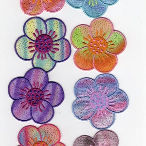 May include: Eight colorful flower patches with embroidered details. Each flower has five petals and a contrasting center. The patches feature a variety of colors, including pink, orange, blue, green, and purple, with glitter accents.