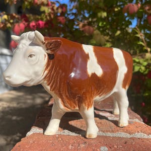 May include: A ceramic cow figurine featuring a glossy white and reddish-brown coat with distinct patches. The animal is depicted in a standing position, showcasing detailed facial features, small horns, and a smooth, reflective surface finish.
