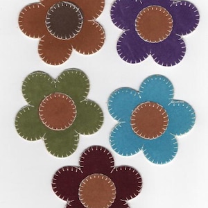 Puede incluir: Cinco formas de flores de fieltro en varios colores: marrón, morado, verde, azul y burdeos. Cada flor tiene un centro marrón contrastante y bordes cosidos en blanco. Estas flores decorativas son adecuadas para manualidades y adornos.