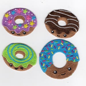 Może przedstawiać: Cztery haftowane naszywki w kształcie pączków z uśmiechniętymi twarzami. Pączki mają różne kolorowe polewy: fioletową z posypką w kształcie gwiazdek, czekoladową z białymi paskami, zieloną z niebieskim wirkiem i niebieską z tęczową posypką.