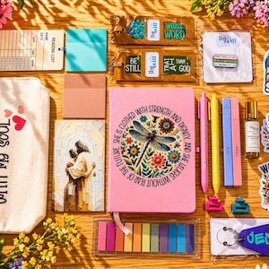 May include: A collection of stationery and inspirational items, including a pink journal with a dragonfly design, pens, stickers, and a keychain that says "JESUS". The items are arranged on a wooden surface.