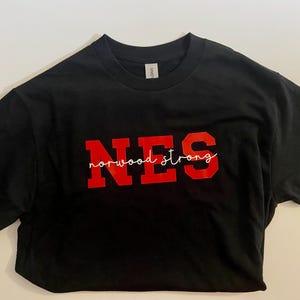 Puede incluir: Camiseta negra con las letras rojas "NES" y las palabras "norwood strong" en una fuente blanca. La camiseta está hecha de un material suave y es adecuada para ropa casual.