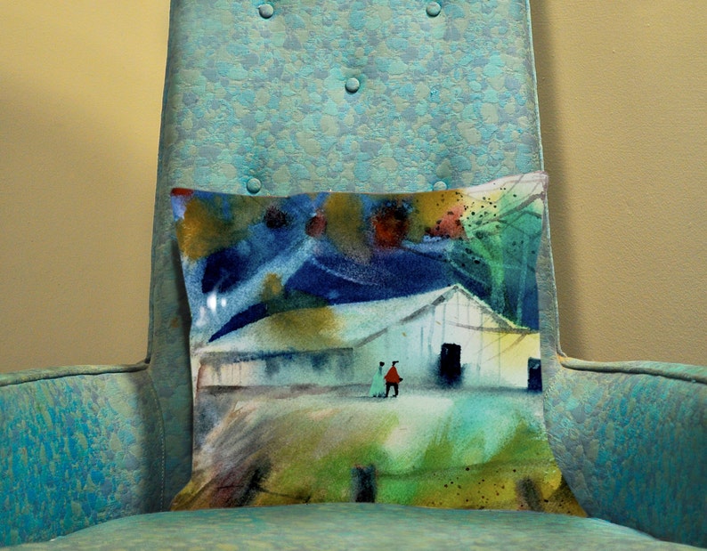 May include: A decorative pillow with an abstract painting of a white building, two figures, and a colorful landscape. The pillow is rectangular and rests on a teal armchair.