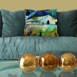 May include: A teal velvet sofa with three gold decorative spheres on a glass table. A decorative pillow with a watercolor painting of a white barn and figures is on the sofa. The painting has blue, green, and red accents.
