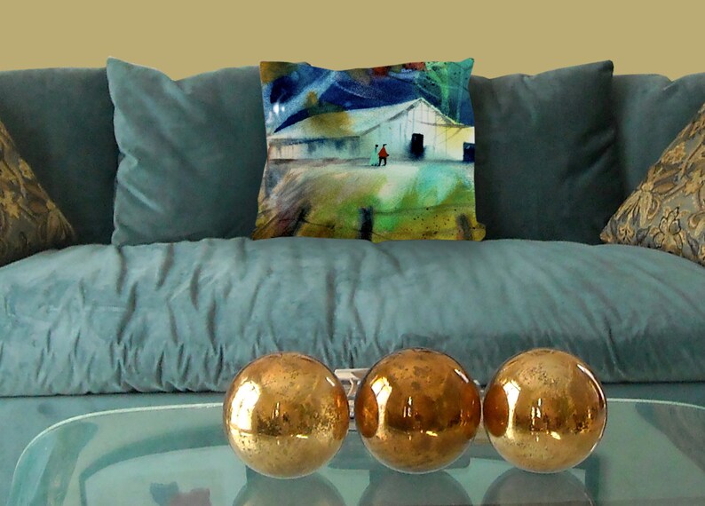 May include: A teal velvet sofa with three gold decorative spheres on a glass table. A decorative pillow with a watercolor painting of a white barn and figures is on the sofa. The painting has blue, green, and red accents.