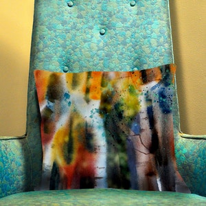 May include: A rectangular decorative pillow with an abstract watercolor design. The pillow features a blend of orange, yellow, blue, and brown hues, with paint splatters. It rests on a teal armchair with button detailing.