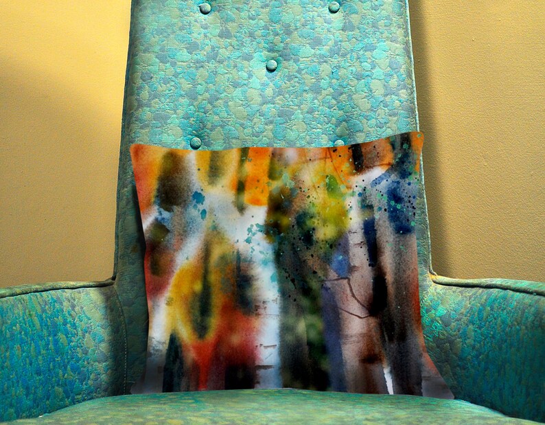 May include: A rectangular decorative pillow with an abstract watercolor design. The pillow features a blend of orange, yellow, blue, and brown hues, with paint splatters. It rests on a teal armchair with button detailing.