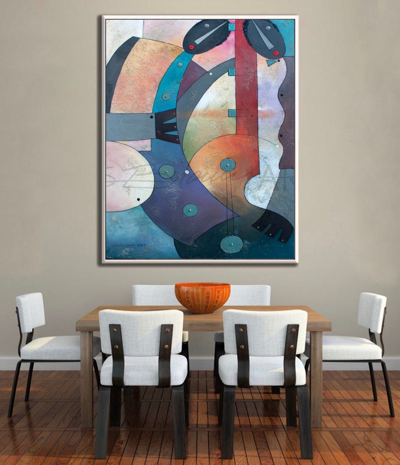 May include: Abstract art painting with geometric shapes in shades of blue, green, orange, and red. The painting is framed in a white frame and hangs on a wall above a wooden table with white chairs.