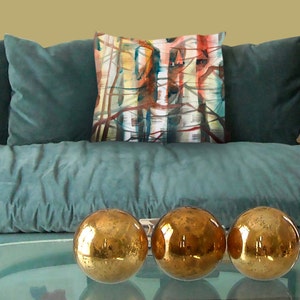 K&ouml;nnte beinhalten: Ein t&uuml;rkisfarbenes Sofa mit drei goldenen Dekorationskugeln auf einem Glastisch. Ein dekoratives Kissen mit einem abstrakten Design in Rot, Blau und Gelb befindet sich auf dem Sofa. Zwei weitere Kissen mit gemusterten Designs sind ebenfalls sichtbar.