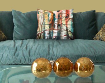 Op de afbeelding: Een turquoise bank met drie gouden decoratieve bollen op een glazen tafel. Een decoratief kussen met een abstract ontwerp in rood, blauw en geel ligt op de bank. Twee extra kussens met patronen zijn ook zichtbaar.