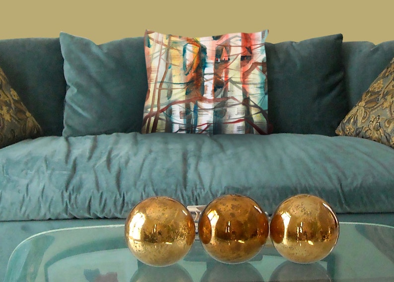 K&ouml;nnte beinhalten: Ein t&uuml;rkisfarbenes Sofa mit drei goldenen Dekorationskugeln auf einem Glastisch. Ein dekoratives Kissen mit einem abstrakten Design in Rot, Blau und Gelb befindet sich auf dem Sofa. Zwei weitere Kissen mit gemusterten Designs sind ebenfalls sichtbar.