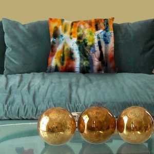 May include: A teal velvet sofa with a decorative pillow featuring an abstract watercolor design in orange, yellow, blue, and black. Three gold metallic spheres sit on a glass table in front of the sofa.
