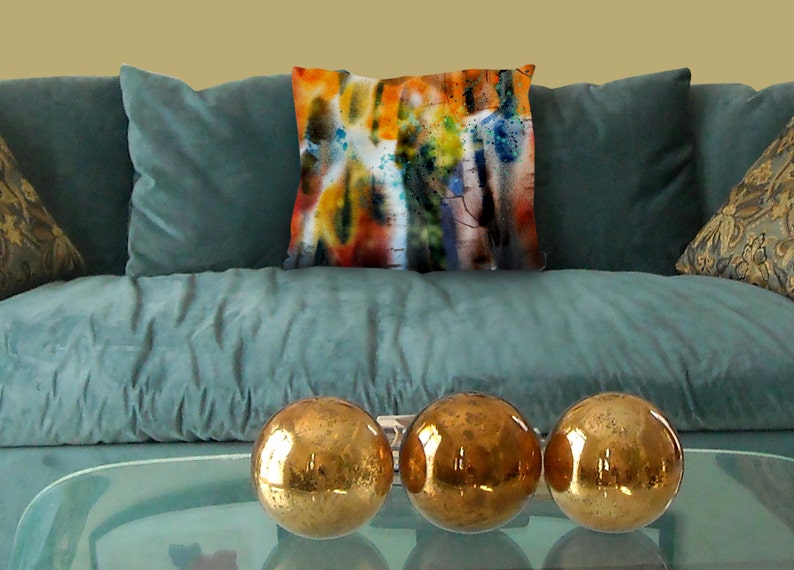 May include: A teal velvet sofa with a decorative pillow featuring an abstract watercolor design in orange, yellow, blue, and black. Three gold metallic spheres sit on a glass table in front of the sofa.