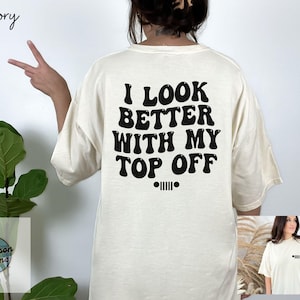 May include: Ivory-colored t-shirt with the text "I LOOK BETTER WITH MY TOP OFF" in black, along with a Jeep logo. The shirt is a relaxed fit.