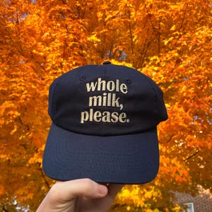 Pode incluir: Um boné de beisebol azul marinho com o texto "whole milk, please." bordado em dourado na frente.