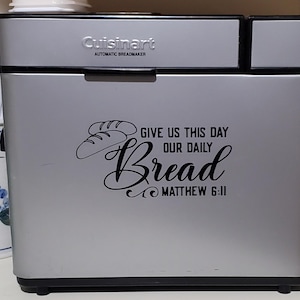 Könnte beinhalten: Ein silberner Cuisinart-Brotbackautomat mit einem schwarzen Vinyl-Aufkleber mit der Aufschrift "Gib uns heute unser tägliches Brot Matthäus 6:11".