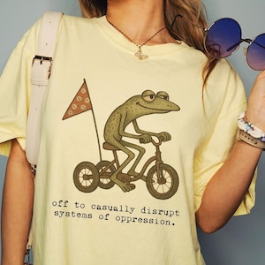May include: Pale yellow t-shirt featuring a cartoon frog on a bicycle with a flag. The text on the shirt says "off to casually disrupt systems of oppression." The shirt is from Banana Comfort Colors.
