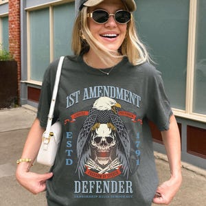 May include: Dark grey t-shirt with a graphic featuring an eagle, skull, and the text "1st Amendment Defender." The shirt also includes the text "Freedom of Speech" and "Censorship Kills Democracy." The shirt is part of the "Comfort Colors" collection.