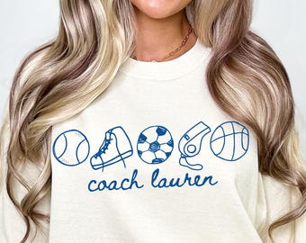 May include: A white t-shirt with a blue graphic of sports equipment and the text "coach lauren" on it.