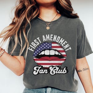 May include: Dark grey t-shirt with the text "FIRST AMENDMENT Fan Club" above a graphic of lips with an American flag design. The shirt is a comfort colors brand.