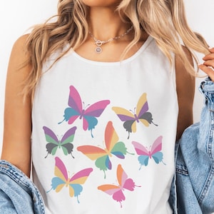 May include: White tank top featuring a colorful butterfly graphic. The butterflies have a variety of colors, including pink, purple, yellow, green, and orange. The tank top is paired with denim shorts and a denim jacket.
