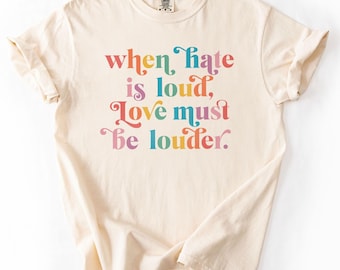 Pode incluir: T-shirt cor marfim com a frase "when hate is loud, Love must be louder" em letras coloridas de estilo retro. A t-shirt é feita de um material macio e confortável, perfeito para uso casual. As palavras "Comfort Colors" estão impressas no canto superior direito.