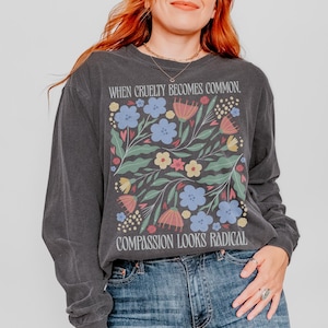 May include: Dark gray long-sleeve shirt with a floral design and the text "WHEN CRUELTY BECOMES COMMON, COMPASSION LOOKS RADICAL." The design includes blue, pink, and yellow flowers. The brand name "pepper COMFORT COLORS" is at the bottom left.