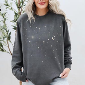 May include: Dark grey long-sleeve sweatshirt with a celestial design. The design features a crescent moon and numerous stars in varying sizes and brightness. The text "pepper COMFORT COLORS" is printed at the bottom left.