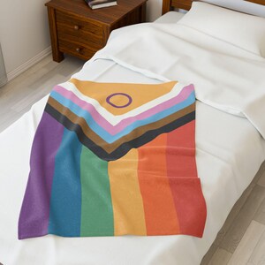 May include: A soft, colorful throw blanket featuring a rainbow design with a purple circle in the center. The blanket is folded and placed on a white bed.