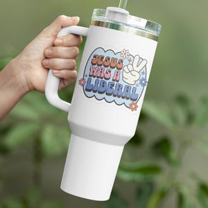 May include: White stainless steel tumbler with a handle and a straw. The tumbler has a sticker with the text "JESUS WAS A LIBERAL" in a retro font with a peace sign and flowers.