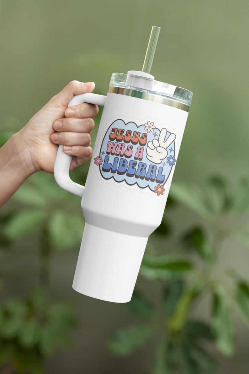 May include: White stainless steel tumbler with a handle and a straw. The tumbler has a sticker with the text "JESUS WAS A LIBERAL" in a retro font with a peace sign and flowers.