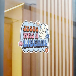 May include: A blue and pink sticker with the text "JESUS WAS A LIBERAL" in a retro font. A peace sign with a white hand is on the right side of the sticker. There are small pink and white flowers around the sticker.