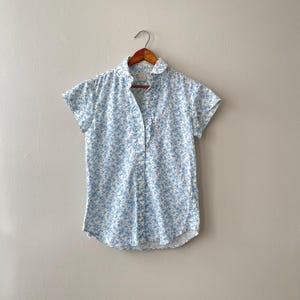 May include: A short-sleeved, button-down shirt with a light blue floral pattern on a white background. The shirt is hanging on a wooden hanger against a neutral wall. The collar is rounded and the shirt has a relaxed fit.