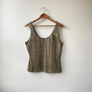 Puede incluir: Una camiseta sin mangas con cuello redondo, con un patrón brillante en oro y negro. La prenda cuelga de una percha de madera contra una pared blanca lisa. La tela tiene un aspecto texturizado.