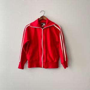 以下が含まれることがあります： 白いストライプと白いジッパーが付いた、赤いジップアップトラックジャケット。 ジャケットは、無地の壁に木製のハンガーに吊るされています。 ジャケットはハイカラーです。