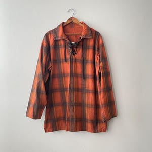 May include: A long-sleeve shirt featuring an orange and dark brown plaid pattern. The shirt has a fold-over collar and a front placket with dark brown laces that tie at the neckline. The garment is displayed on a wooden hanger against a plain wall.