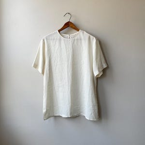 May include: Off-white short-sleeve top hanging on a wooden hanger. The top has a round neckline and a simple, classic design. The fabric appears to be lightweight and slightly textured, with a subtle sheen.