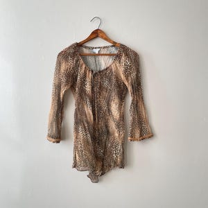 May include: A sheer, long-sleeved top with a brown and tan leopard print pattern. The top has a scoop neckline and is hanging on a wooden hanger. The sleeves have a slight bell shape and the fabric appears lightweight.