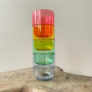 May include: A stack of five colourful, transparent drinking glasses. The glasses are arranged in a vertical column, with each glass a different colour: pink, orange, yellow, green, and blue. The glasses are sitting on a wooden surface.