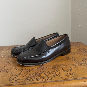 May include: A pair of black leather penny loafers. The shoes have a classic design with a stitched vamp and a low heel. The loafers are displayed on a wooden surface.