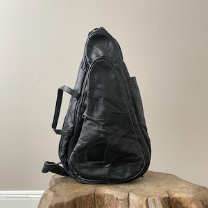 以下が含まれることがあります： パッチワークデザインの黒い革製バックパック。バッグは丸みを帯びた形状で、上部にハンドルとショルダーストラップが付いています。木製の表面に置かれています。