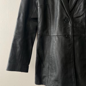 May include: A black leather blazer with a classic design. The blazer features a notched lapel, two-button closure, and long sleeves. The leather appears to be smooth and has a slight sheen. The garment is tailored with seams for a structured fit.